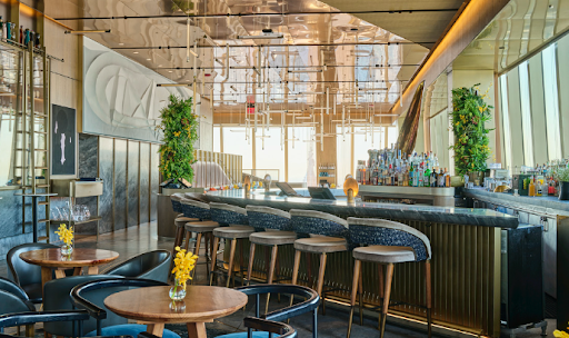 Modern, upscale bar with curved counter, high stools, round tables with yellow flowers, large windows, and decorative lighting in a brightly lit space.