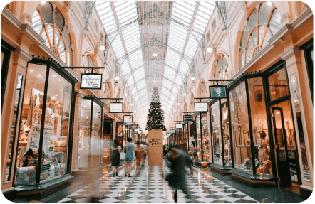 A grand, well-lit shopping arcade with a glass ceiling adorned with festive decorations, including a large Christmas tree at its center. Shoppers in motion blur walk along the checkered floor, flanked by various stores on either side displaying an array of goods in their windows.