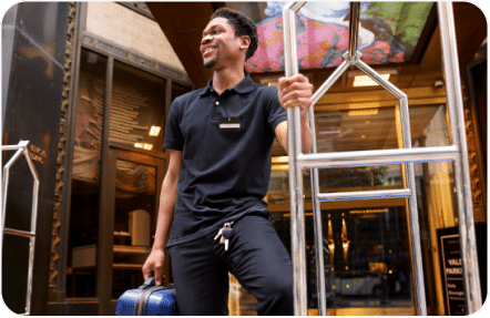 A smiling hotel staff member, dressed in a black uniform, stands at the entrance of a hotel. He is holding a blue suitcase in his right hand and has keys clipped to his waistband. The hotel door and a golden luggage cart are visible in the background.