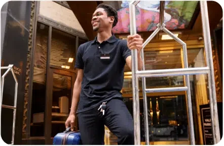 A hotel staff member in a black uniform stands by a luggage cart, holding a small blue suitcase, smiling, with a building entrance in the background.