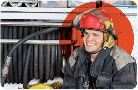 A firefighter, wearing protective gear and a red helmet, is smiling while seated. Behind him, there are coiled fire hoses and equipment stored in a fire truck. The background includes a panel with a metal diamond plate texture, partially covered by a red circle graphic.