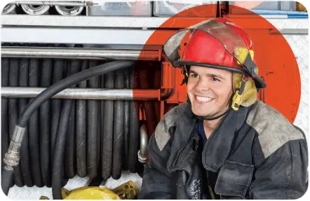 A firefighter wearing a red helmet and protective gear smiles, seated in front of coiled hoses on a fire truck.