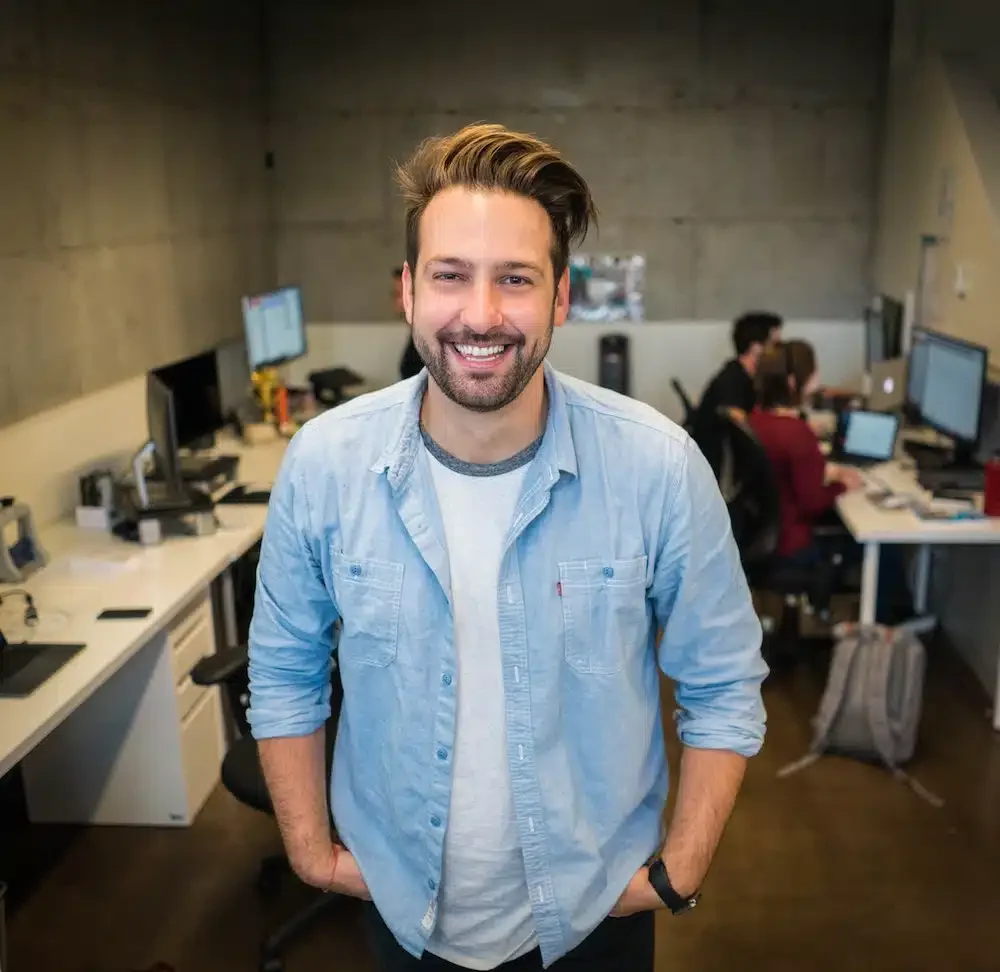 A man with a beard and casual attire stands smiling in an office with three other people working at desks with computers.