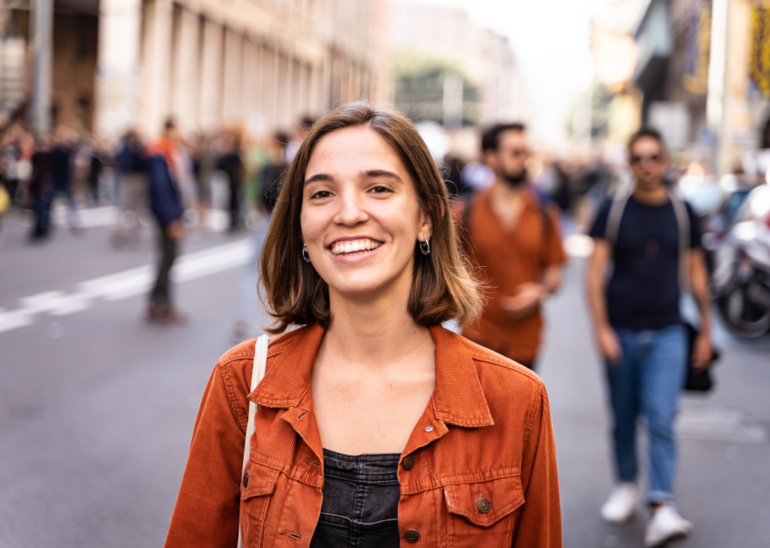 A young woman in an orange jacket smiles at the camera while standing on a busy city street with people in the background.