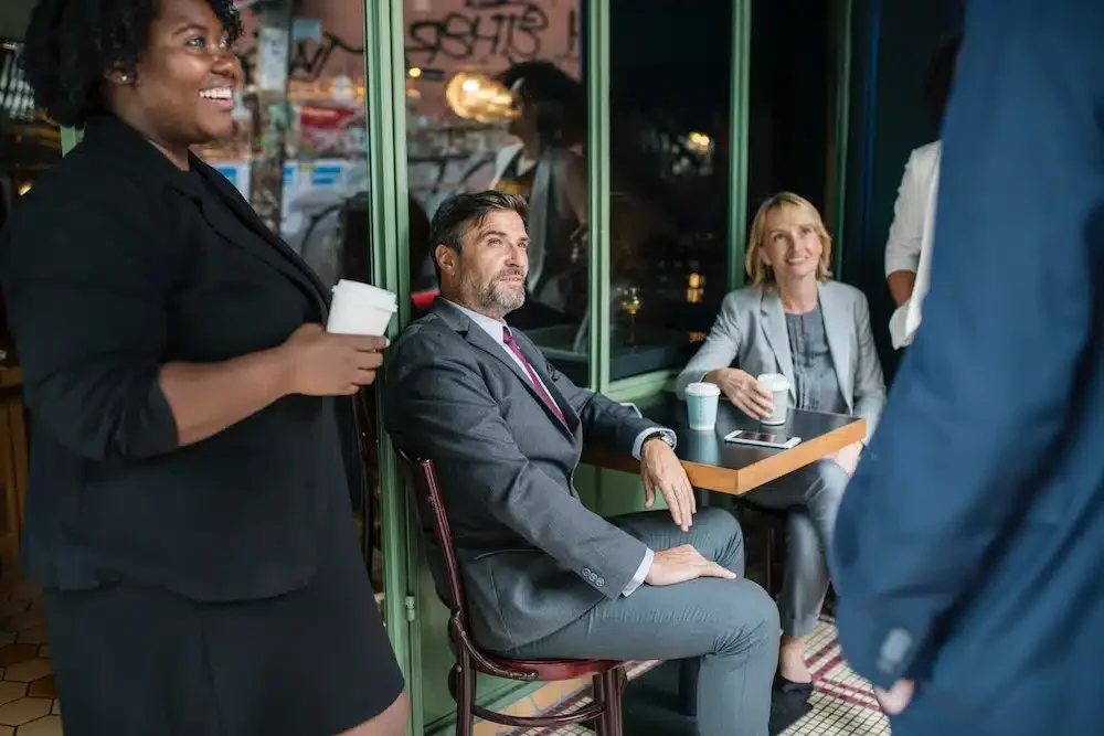 A group of people in business attire are chatting and drinking coffee inside a cafe. One person is sitting at a table while others stand around and smile.