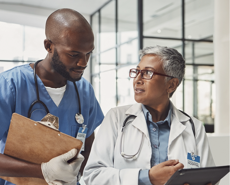 Two healthcare professionals, one in blue scrubs holding a clipboard and one in a white coat with a stethoscope, are discussing patient information in a hospital setting.
