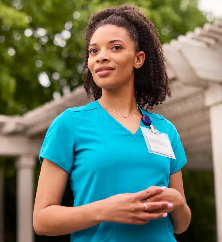 A person in a turquoise medical uniform with a name badge stands outdoors, with trees and a pergola in the background.