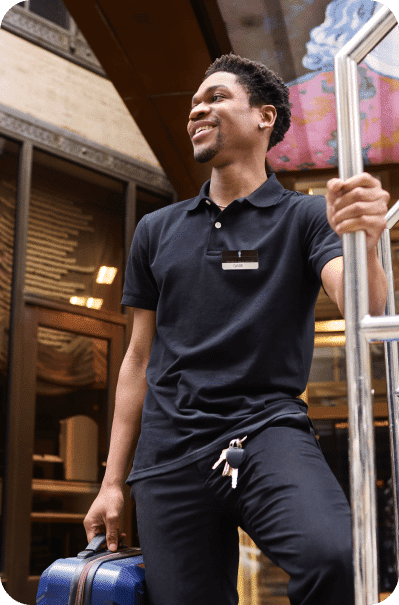 Smiling man in a black polo shirt with a name tag, holding a luggage handle in one hand and a suitcase in the other. He is standing outside a building with glass doors. Keys are attached to a keyring hanging from his pocket. He looks cheerful and ready to assist.