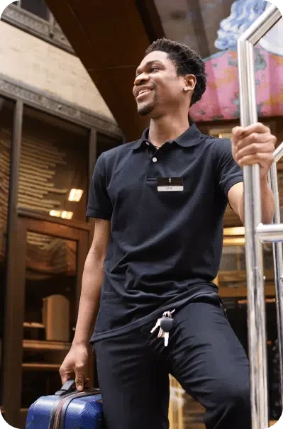 A man dressed in a black polo shirt, holding onto a trolley cart with one hand and a suitcase in the other, smiles while standing outside a building.