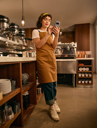 A woman wearing an apron stands in a coffee shop using her smartphone, with coffee machines and shelves of cups visible around her.