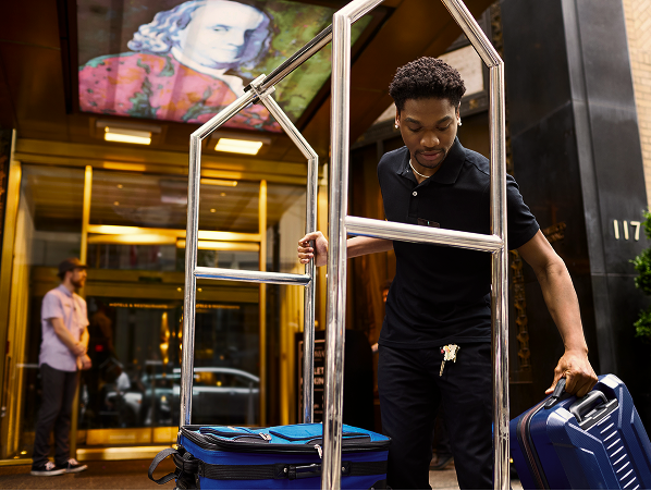 A hotel employee moves luggage with a trolley outside a building entrance; a portrait of Benjamin Franklin is displayed above the door.