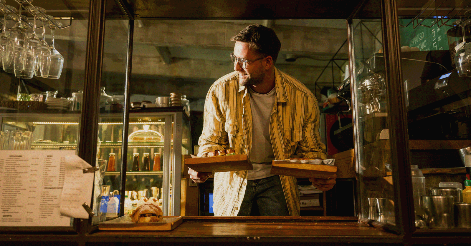 A barista in a yellow striped shirt reaching over a counter to serve two wooden trays of food.