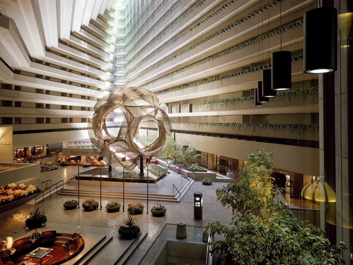 Spacious modern hotel atrium with a large geometric sculpture in the center, surrounded by greenery, seating areas, and multiple stories of balconies.