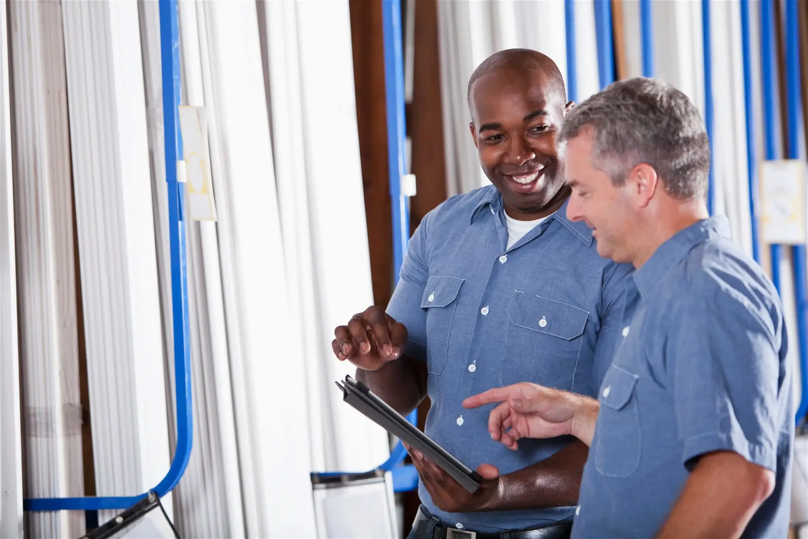 Two men in blue work shirts are discussing something on a tablet while standing in a warehouse aisle with metal pipes in the background.