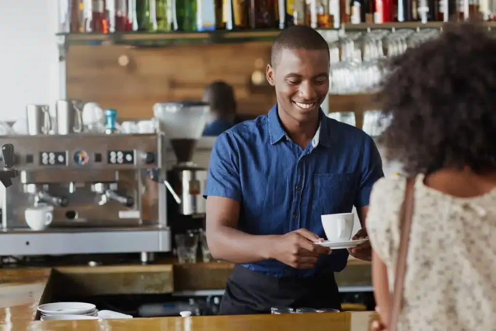 A barista in a blue shirt serves a cup of coffee to a customer at a café counter.