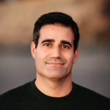A man with short dark hair and a slight smile is standing outdoors, wearing a dark shirt. The background is blurred.