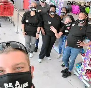 A group of people wearing matching black shirts and face masks with text, standing together in what appears to be a store. One person in the foreground is taking a selfie.