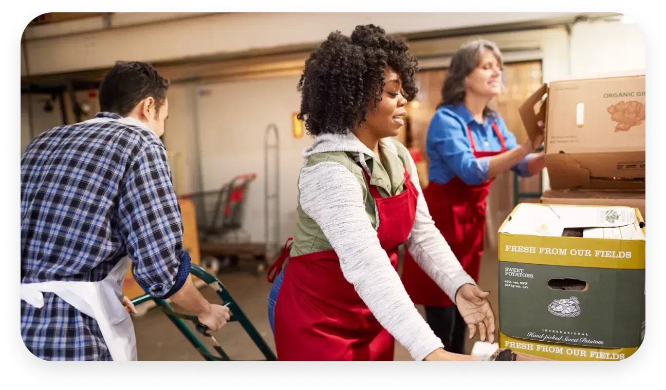 Three people wearing red aprons are working in a warehouse, moving and organizing boxes. Two are stacking boxes, and one is using a pallet jack.