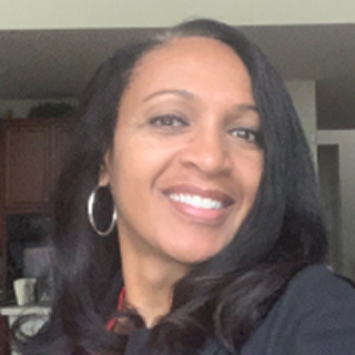Smiling woman with long black hair and hoop earrings, wearing a dark top, standing indoors with a kitchen in the background.