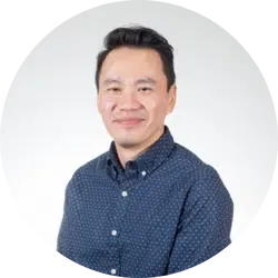 A person with short black hair wearing a dark blue button-up shirt with small white polka dots, smiles at the camera against a light background.