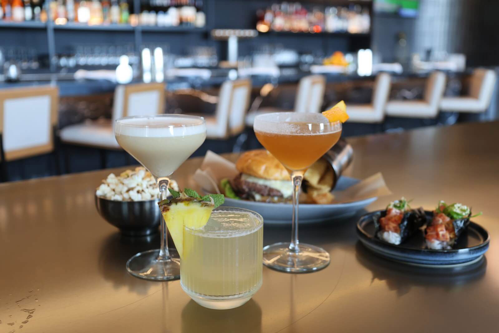 Three cocktails, a bowl of popcorn, a burger with fries, and a plate of sushi are arranged on a bar counter with chairs and shelves in the background.