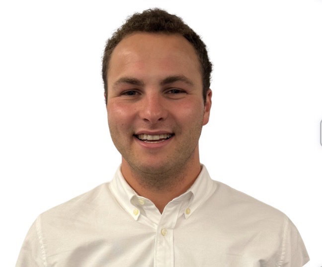 A person with short curly hair, wearing a white button-up shirt, is smiling at the camera against a plain white background.