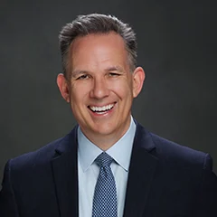 A man in a suit and tie smiles in front of a plain dark background.