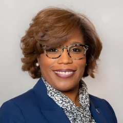 A woman with short brown hair, wearing tortoiseshell glasses, a navy blazer, and a patterned scarf, smiling against a light gray background.