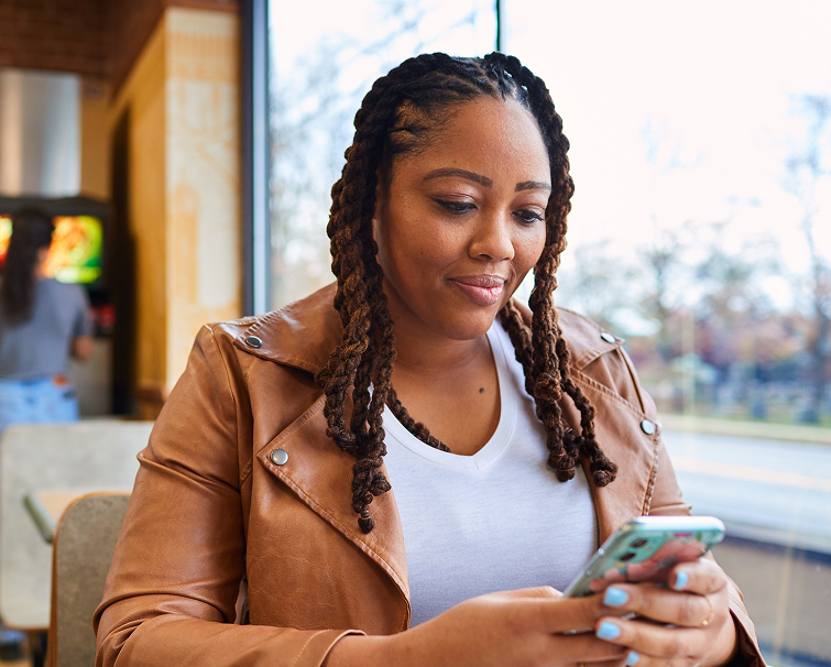 A woman with long braided hair sits at a table in a casual restaurant, looking at her smartphone and smiling slightly.