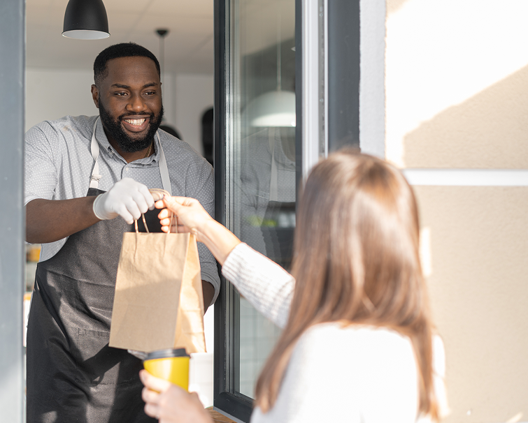 A man wearing an apron and gloves hands a paper bag to a woman holding a cup at a takeout window.