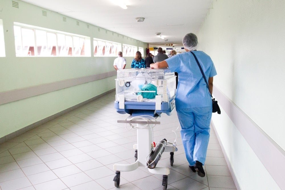 A healthcare worker in blue scrubs pushes a medical cart along a brightly lit hospital corridor, walking away from the camera towards other staff and patients in the distance.