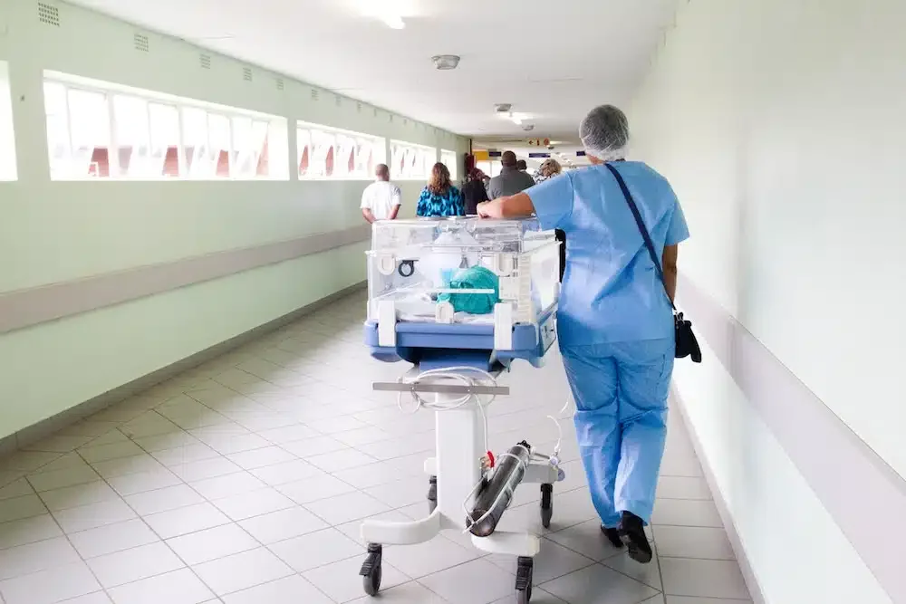 A healthcare worker in blue scrubs pushes a baby incubator along a hospital corridor, with several people visible in the background.