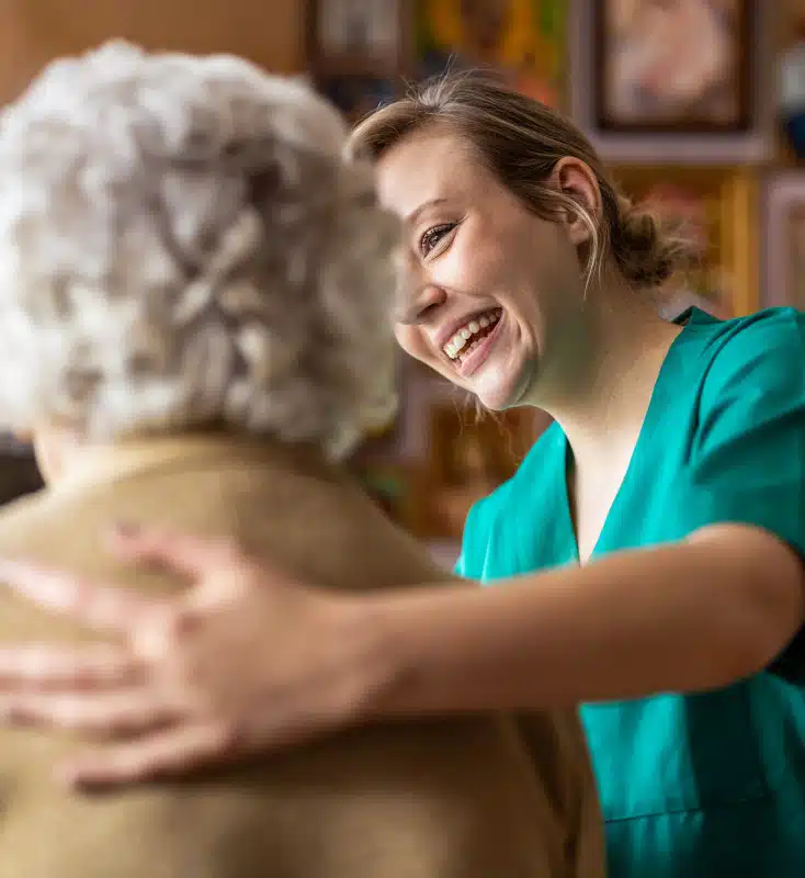A young caregiver in green scrubs smiles while placing her arm around an elderly person with white hair, creating a comforting and positive atmosphere.