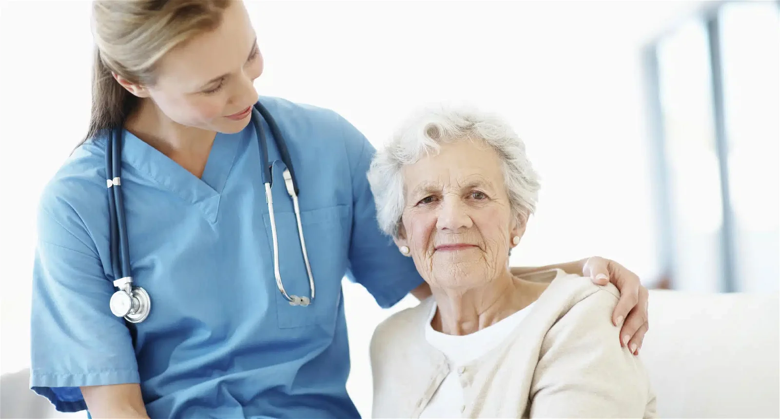 A healthcare worker in blue scrubs with a stethoscope reassures an elderly woman with gray hair, who is wearing a beige cardigan.