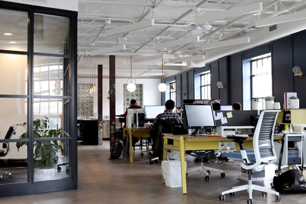 Modern office space with employees working at desks, featuring stylish interior design with exposed brick, large windows, and an industrial ceiling.
