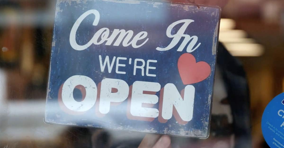 A sign hangs in a store window with the message "Come In We're OPEN" written in white letters on a blue background with a red heart next to the word "OPEN." The sign has a slightly weathered appearance, much like they've embraced on demand pay, always ready for your patronage.