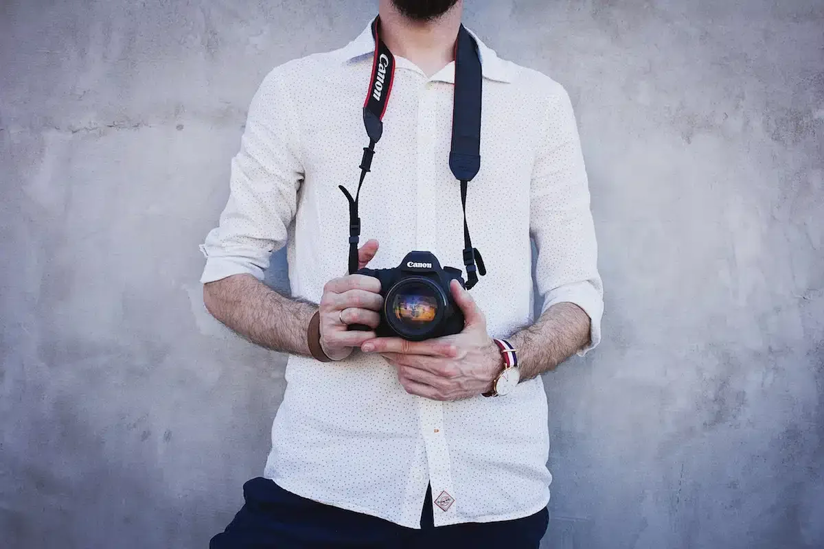 A person wearing a white shirt holds a Canon camera with both hands, against a gray concrete wall background.
