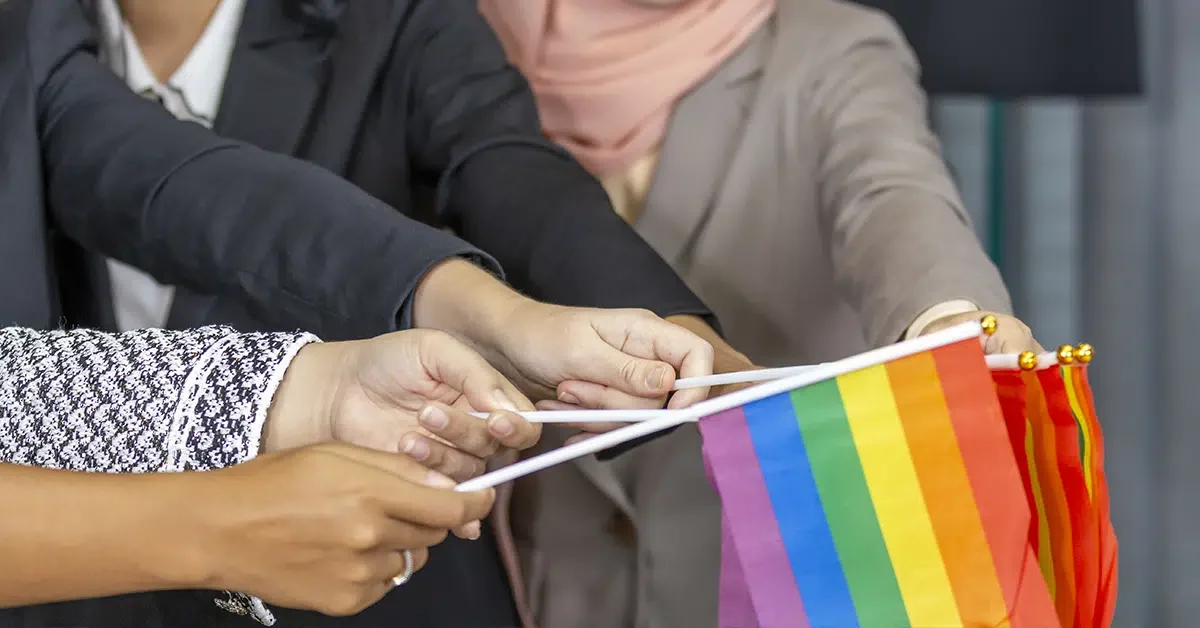 Four people extend their hands, grasping small sticks together. Each stick holds a piece of a rainbow flag, symbolizing LGBTQ+ pride. The individuals are dressed in business attire, including suits and a hijab, signifying a diverse, inclusive environment. The background is blurred.
