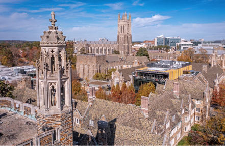 Aerial view of a Gothic-style university campus with multiple stone buildings and a tall church tower, trees with fall colors, and modern buildings in the background under a clear blue sky.