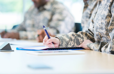 Three individuals in military camouflage uniforms are seated at a table, focused on writing. The person in the foreground is holding a pen and writing on a clipboard. The other two are blurred in the background. The setting appears to be a classroom or meeting room.