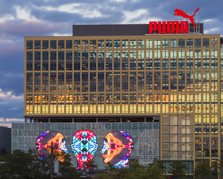 Office building with a red Puma logo on the roof and a colorful illuminated face art installation on the lower facade at sunset.