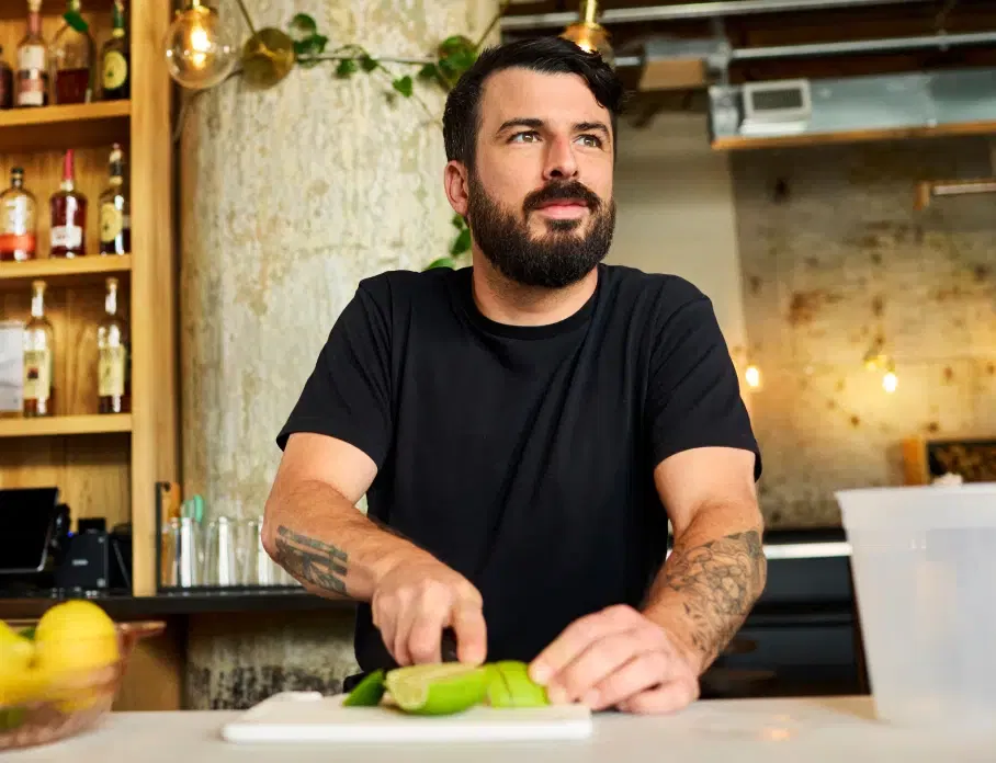 A man with a beard and tattoos chops green vegetables on a white cutting board in a modern kitchen with wooden shelves and various bottles in the background.