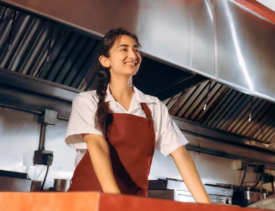 A smiling woman in a white shirt and red apron stands behind a counter in a commercial kitchen with stainless steel equipment in the background.
