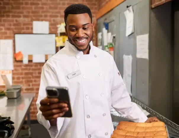 A person in a white chef's coat smiles while looking at a smartphone and holding a tray of freshly baked bread in a kitchen.