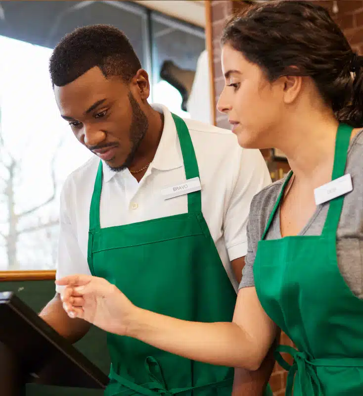 Two café employees in green aprons, one male and one female, are looking at a screen. The woman is pointing at the screen, explaining something to the man.