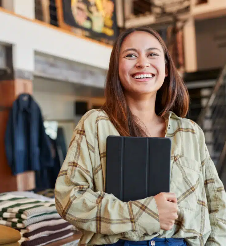 A woman with long hair and a beige plaid shirt is smiling while holding a black tablet. She is standing indoors near a staircase and clothing display.