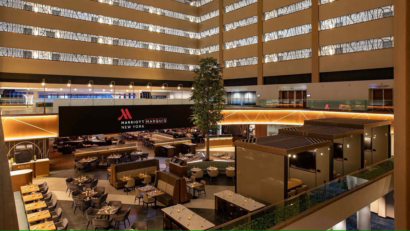 Modern hotel atrium with multiple floors, stylish dining areas, private booths, and a large Marriott Marquis New York sign. Warm lighting and contemporary decor throughout the space.