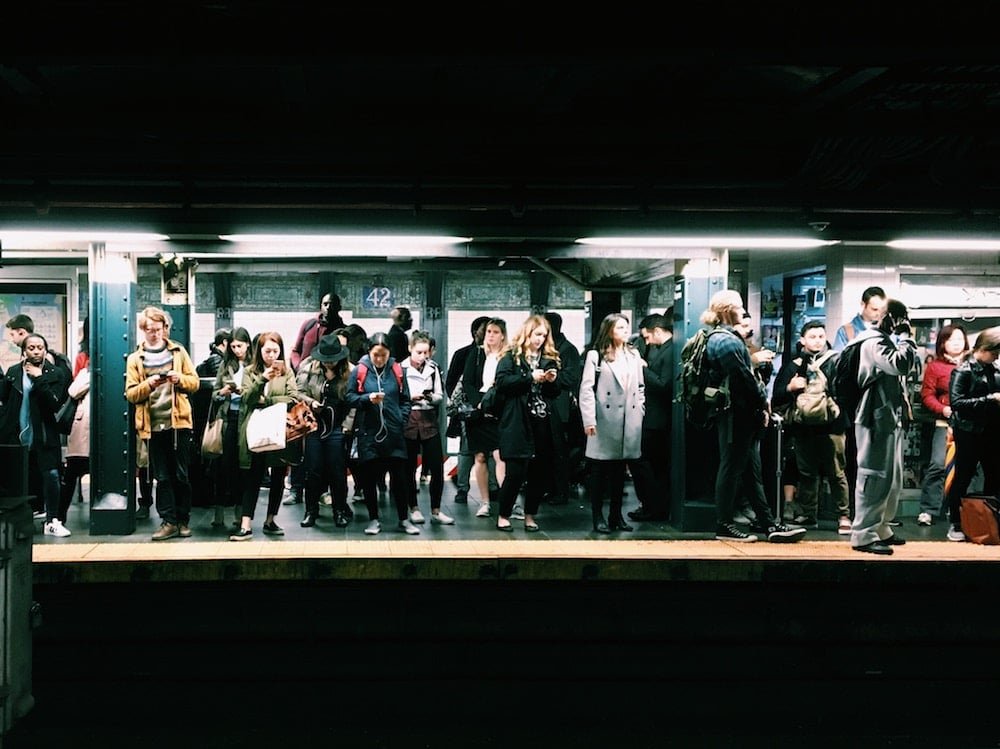 A group of diverse commuters waiting on a subway platform, standing behind the yellow safety line, with some looking at their phones.