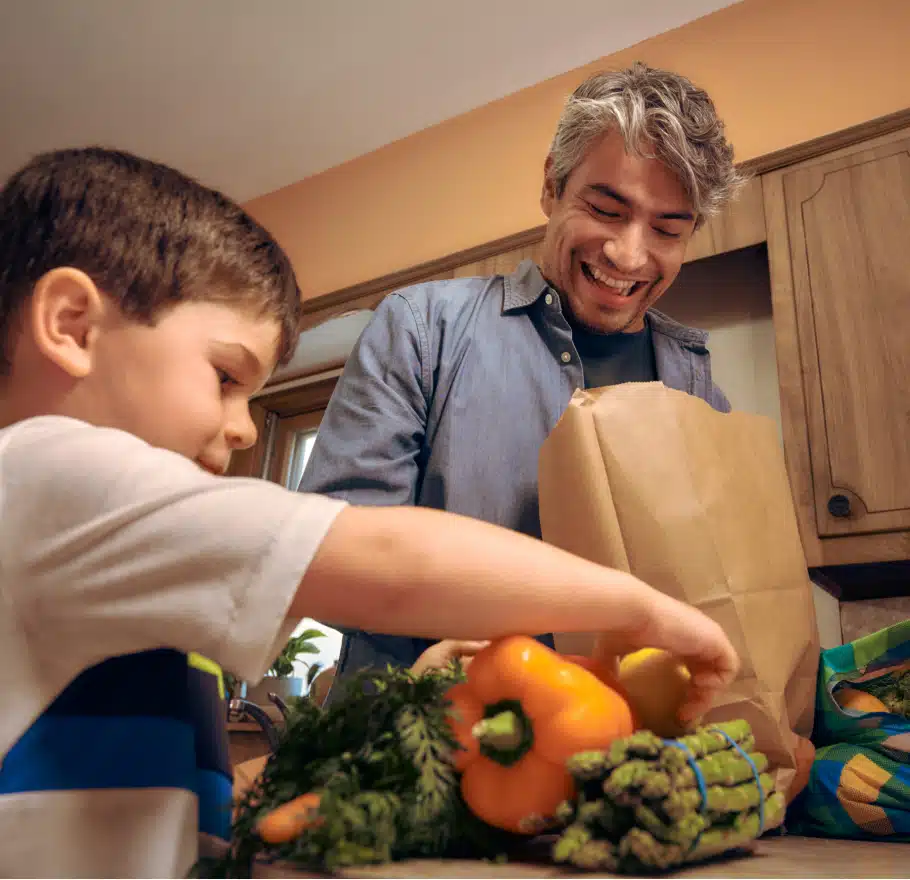 A man and a young boy unpack groceries in a kitchen, with fresh vegetables like bell peppers, asparagus, and carrots spread on the counter.
