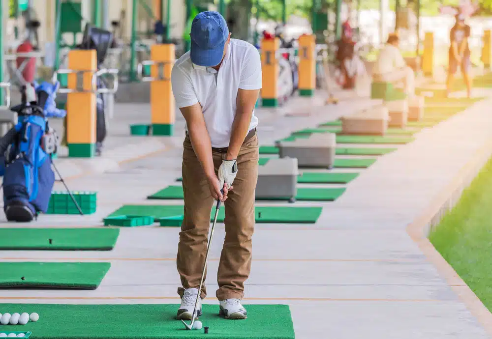 A person dressed in a white shirt, brown pants, and a blue cap practices putting on a golf mat at an indoor golf range.